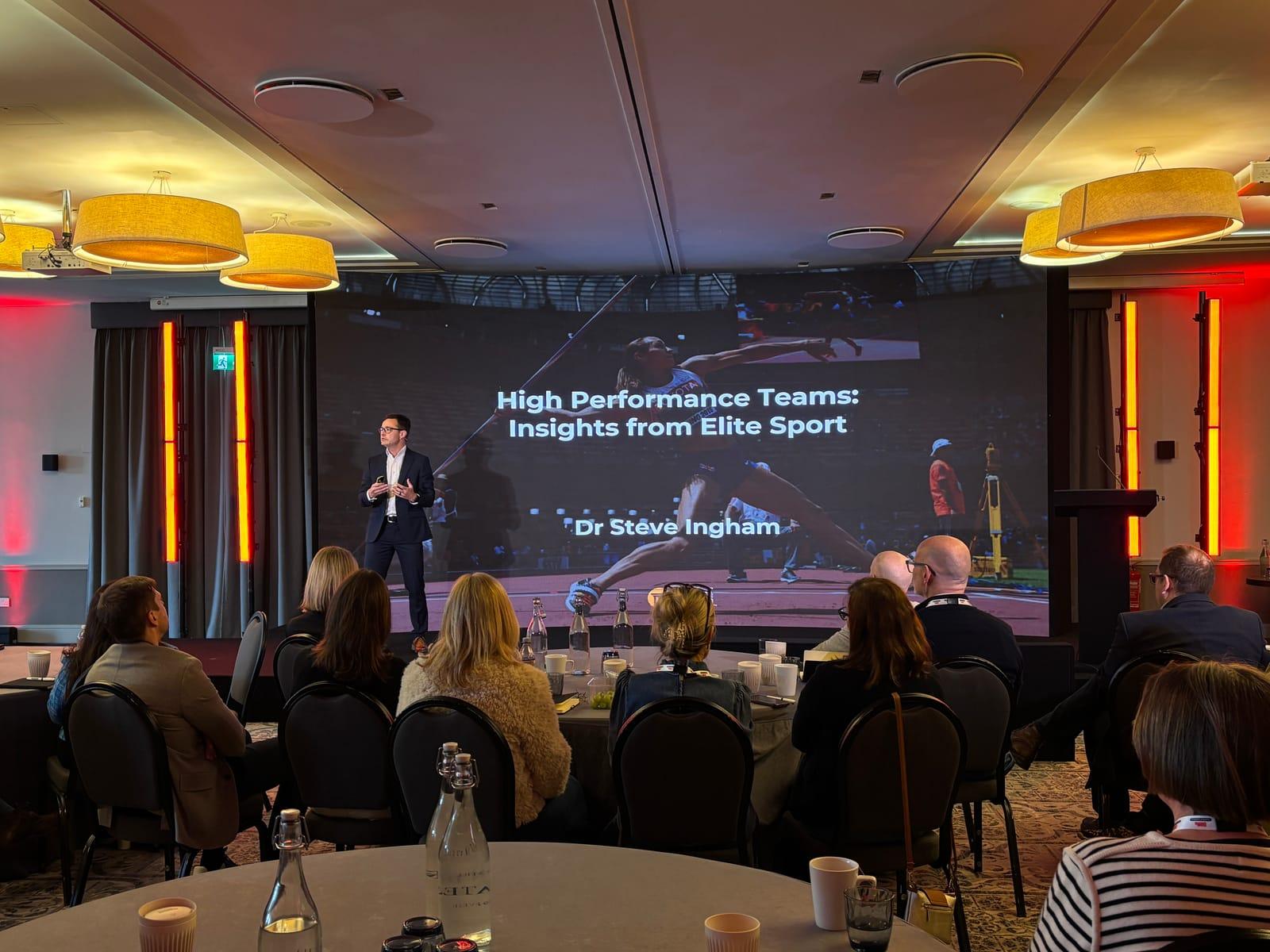 Steve Ingham on stage with audience and athlete imagery on screen